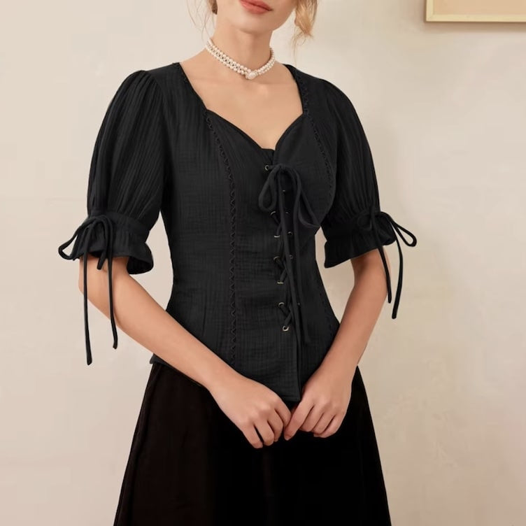 A person wearing a black blouse with lace-up detailing and puff sleeves, paired with a black skirt and a pearl necklace.
