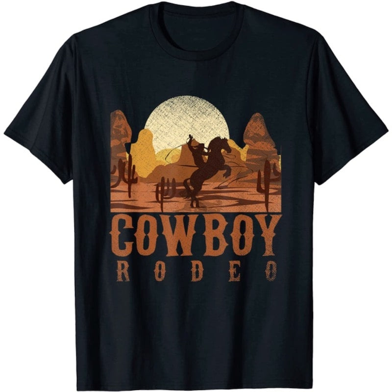 Black t-shirt with "Cowboy Rodeo" graphic featuring a cowboy riding a rearing horse in a desert landscape with cactuses.