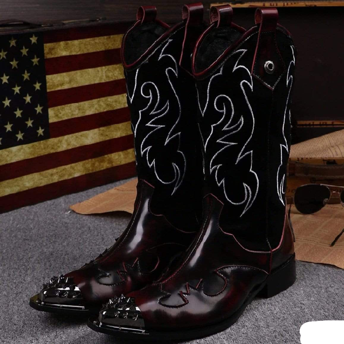 Burgundy cowboy boots with silver toe spikes and white embroidery, presented in front of a vintage American flag backdrop.