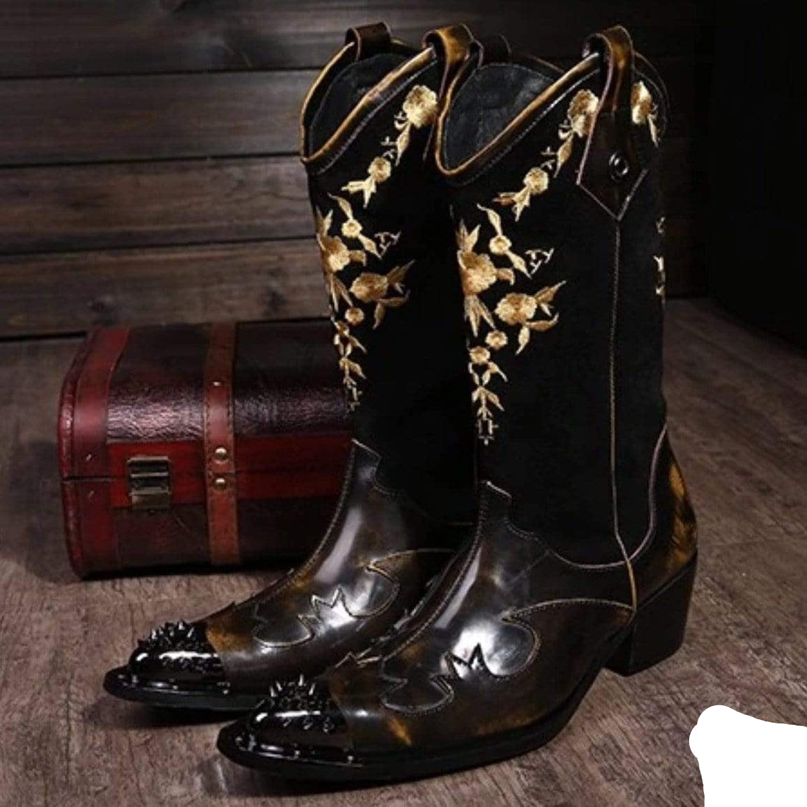 A pair of ornate black and gold cowboy boots with spiky toe caps, positioned on a wooden floor next to a small wooden chest.
