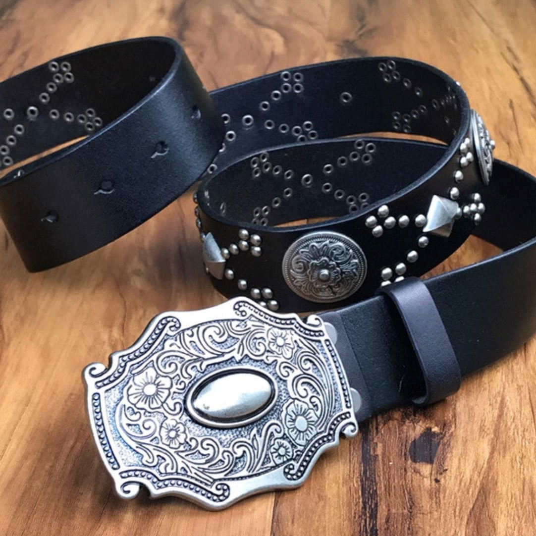 Two black leather belts, one with a decorative silver buckle and the other with silver studs and embellishments, on a wooden surface.