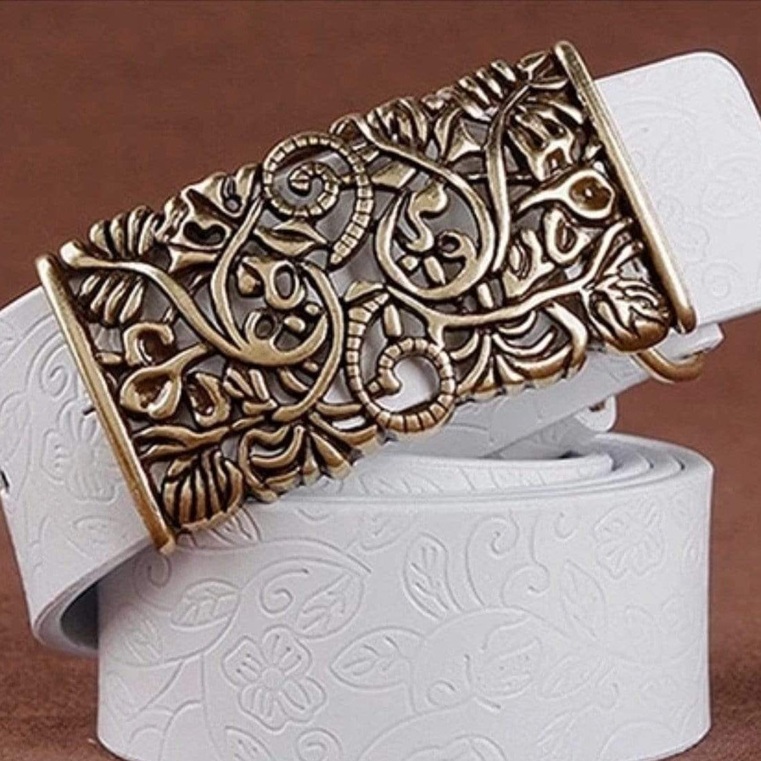 Close-up of a white leather belt with a decorative bronze buckle featuring floral and vine designs, displayed on a brown surface.
