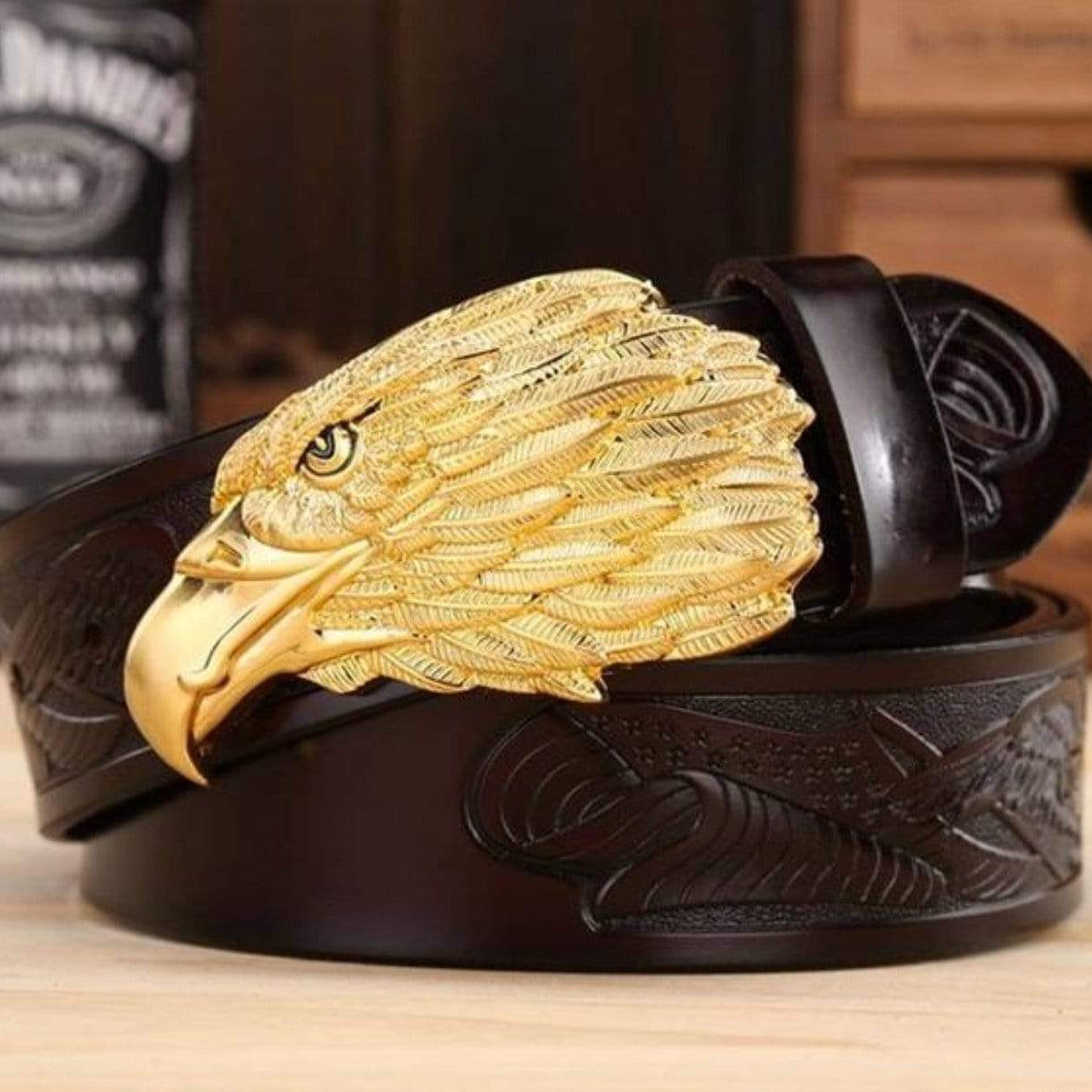 Close-up of a dark brown leather belt with a large gold eagle buckle. The belt has embossed feather details. A bottle of liquor is visible in the background.