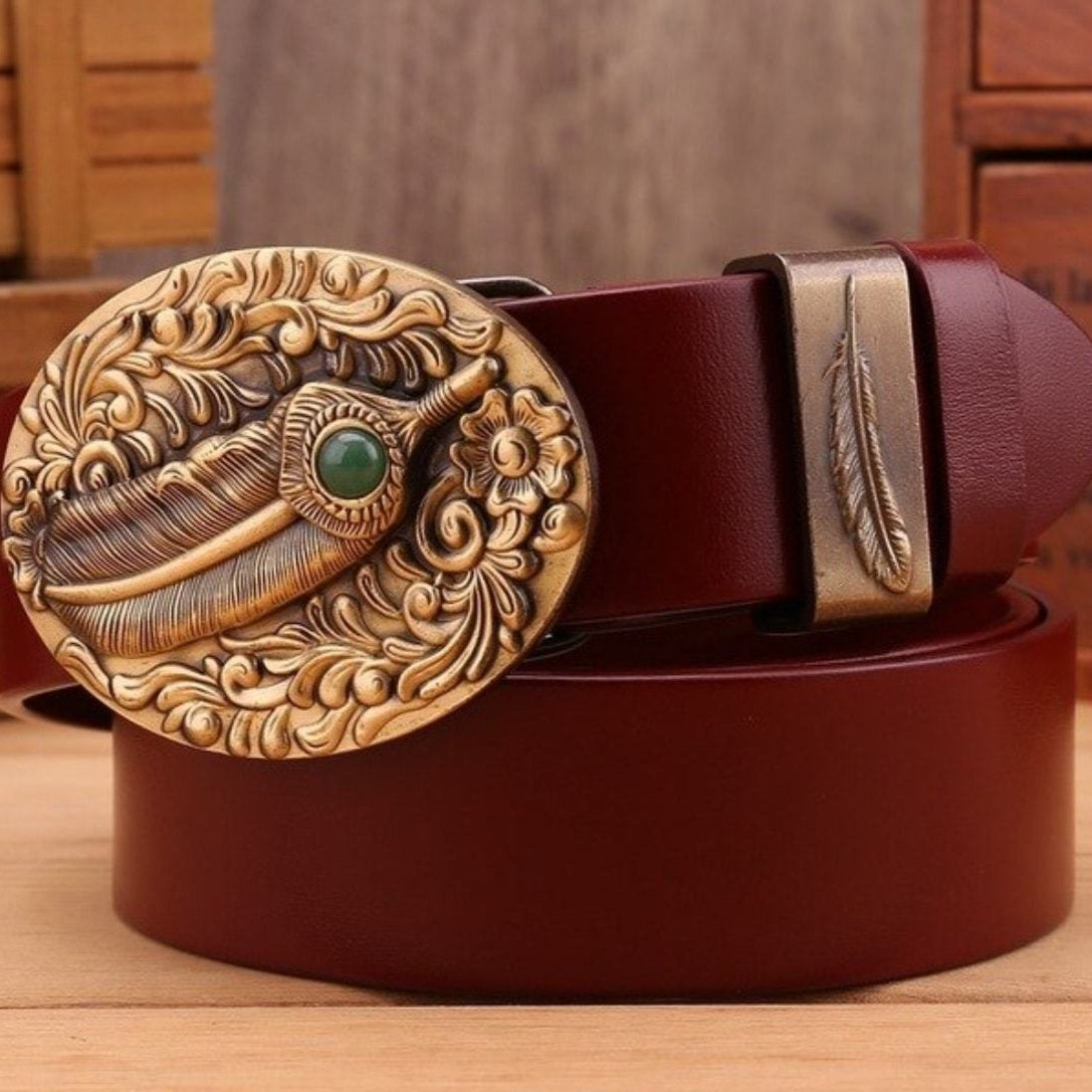 Close-up of a red leather belt with a large, ornate, round brass buckle featuring a feather and green stone detail. The belt also has a matching brass loop with a smaller feather design.
