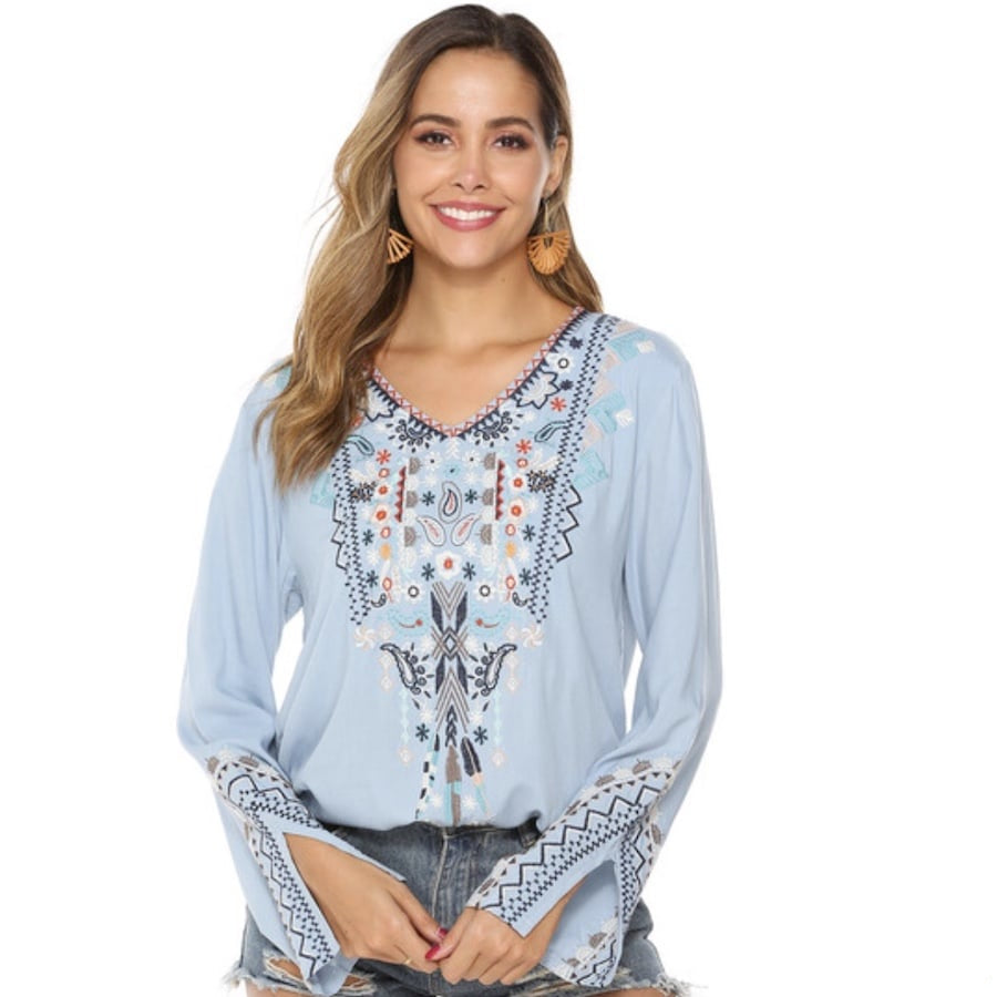 A woman wearing a light blue, embroidered, long-sleeved blouse with denim shorts.