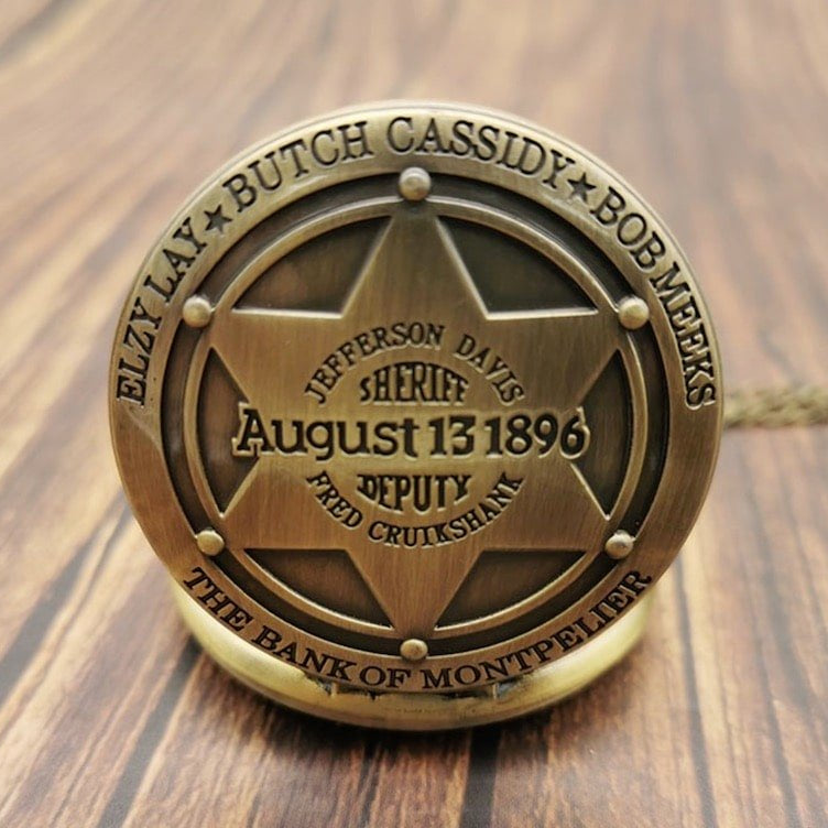 Close-up of a bronze-colored pocket watch with a six-pointed star on its cover, featuring names like "Butch Cassidy," "Jefferson Davis," and "Fred Cruikshank," along with "August 13 1896" and "The Bank of Montpelier" inscribed on it. The watch sits on a wooden surface.