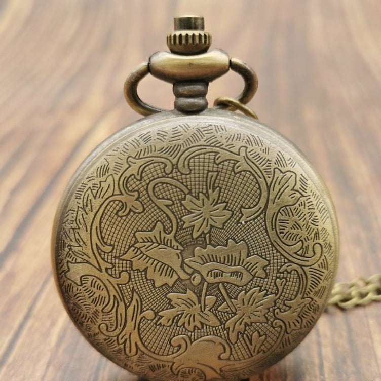 Close-up of a bronze-colored pocket watch with an ornate floral design, resting on a wooden surface.