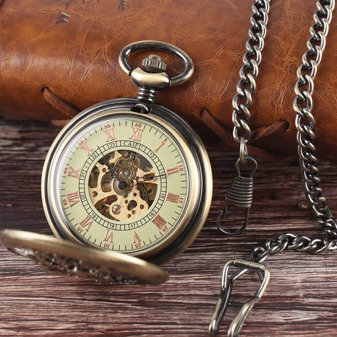 Close-up of a bronze-toned pocket watch with a visible mechanical interior, Roman numerals, and a chain, sitting on a wooden surface with a leather object in the background.