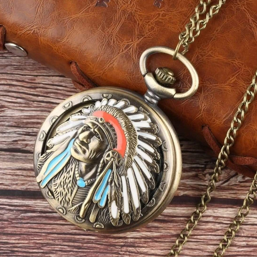 Close-up of a vintage-style pocket watch with a Native American chief design, lying on a brown leather notebook and wooden surface, connected to a gold chain.