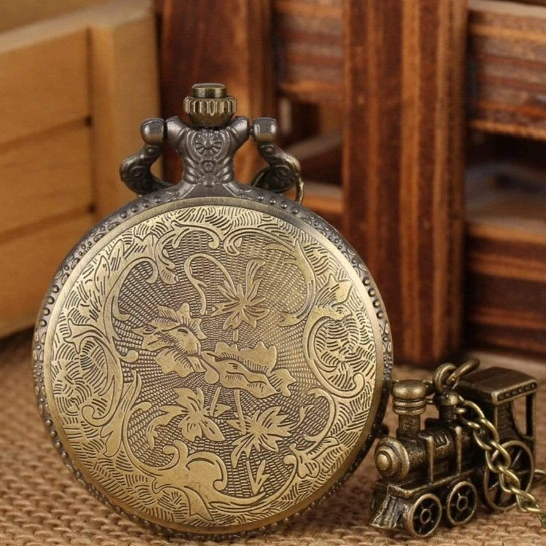 Close-up of an ornate bronze pocket watch and train charm necklace on a textured surface with wooden accents in the background.