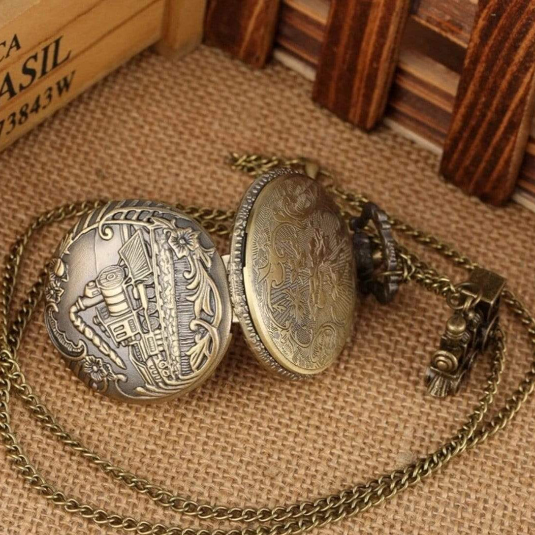 Open antique bronze pocket watch with train-shaped chain on burlap.
