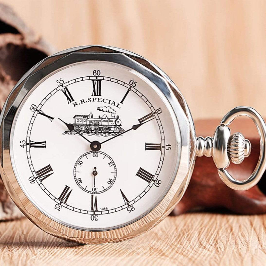 Close-up of a silver pocket watch with a white face, black Roman numerals, and a train illustration at the top, resting on a wooden surface.