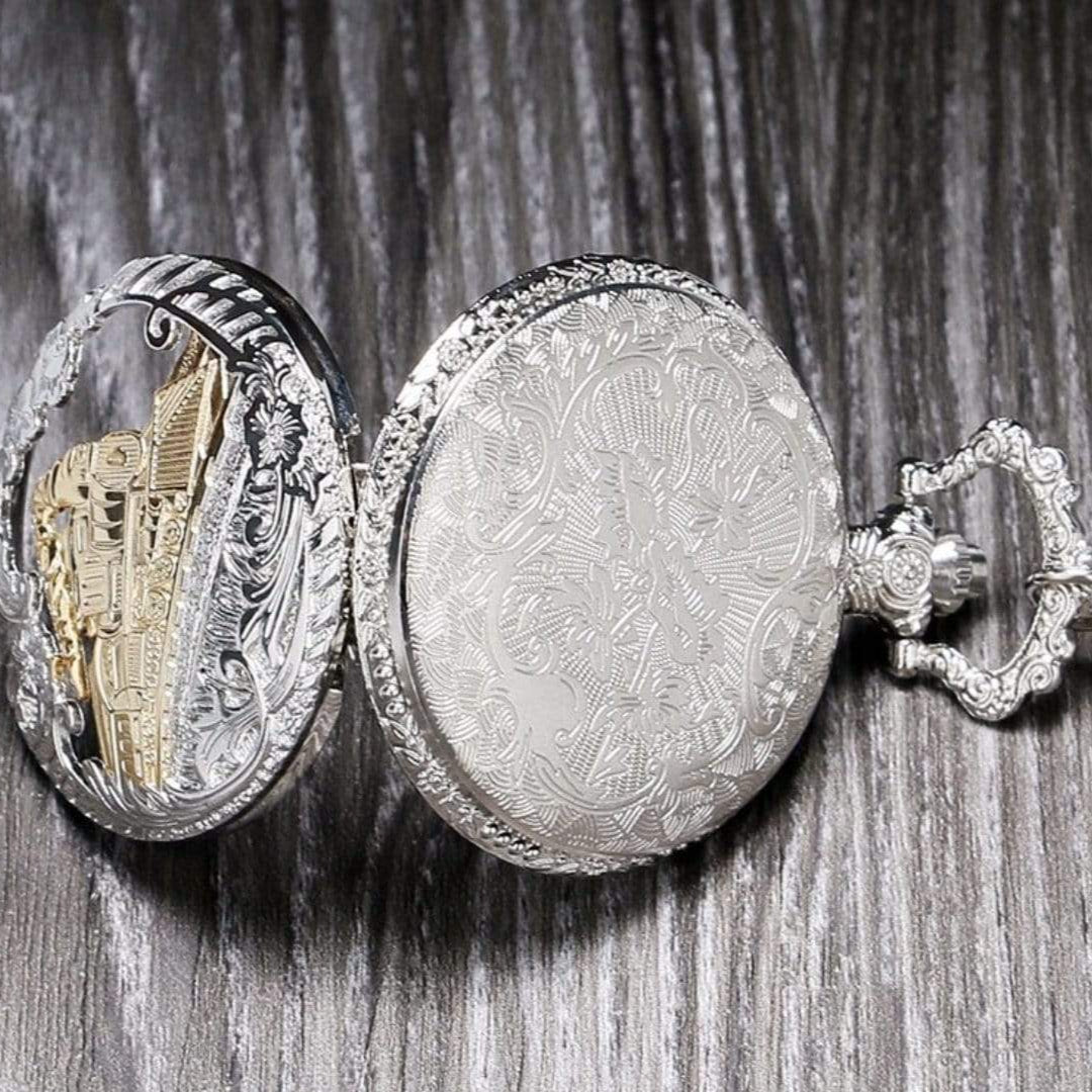 An ornate, silver pocket watch rests on a wooden surface. The watch is open, revealing a gold-colored interior design. 
