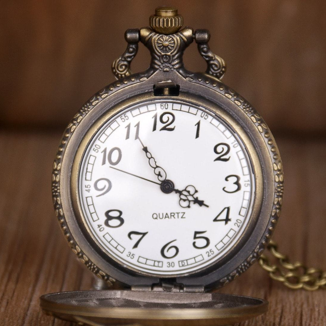 Close-up of an antique-style pocket watch with an open front cover, showcasing a white face with black numbers, a Quartz label, and gold hands. The watch has an ornate metal casing and is placed on a wooden surface with a chain partially visible.
