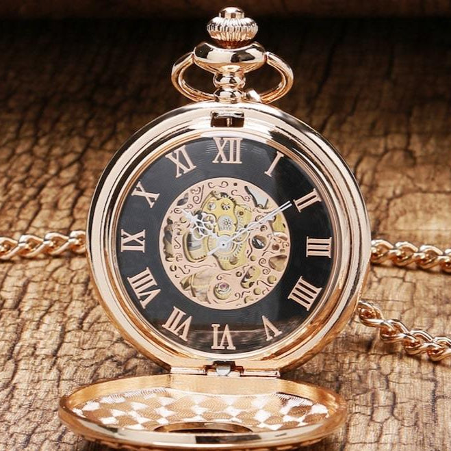 Gold-colored pocket watch with black face and Roman numerals. The watch has a cut-out center showing the gears. It is on a chain and lying on a wooden surface. 
