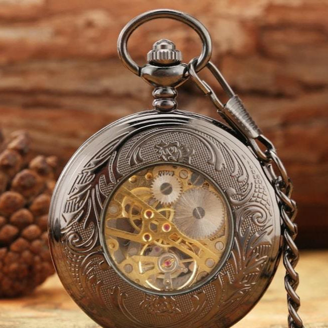 Close-up of an ornate black pocket watch with visible gears and chain against a wooden background.
