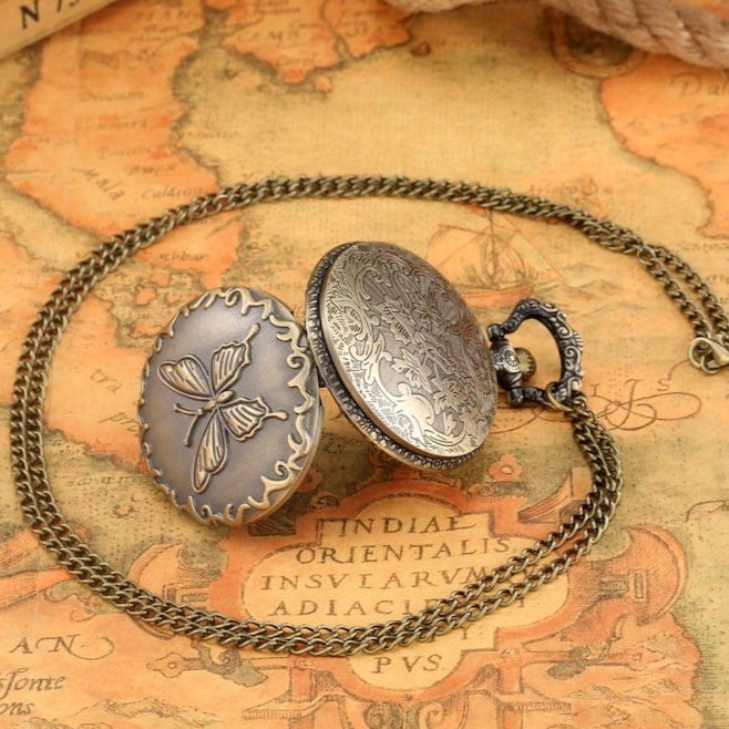 Close-up of an antique, ornate pocket watch necklace with an oval pendant displaying a butterfly, resting on a vintage map background.