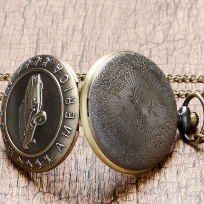 Open bronze pocket watch with a car emblem and chain on a wooden surface.
