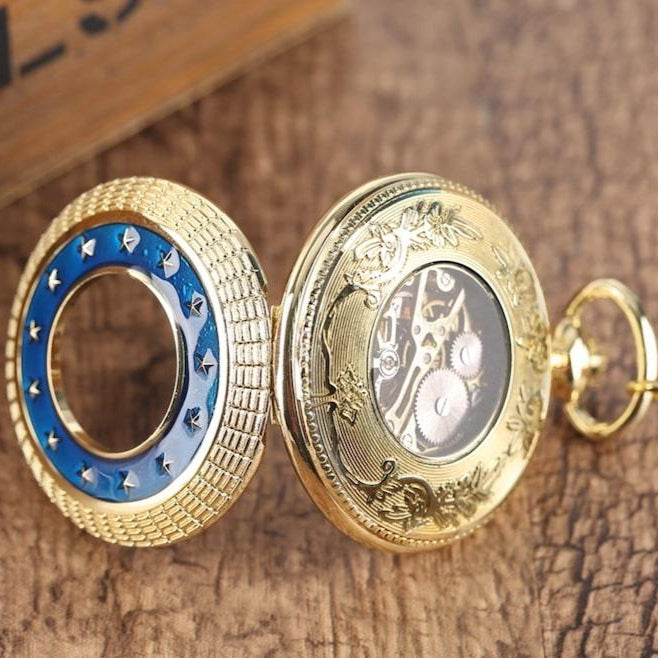 Close-up of an open, golden pocket watch with blue and gold accents, showcasing intricate gears and detailed craftsmanship on a wooden surface.
