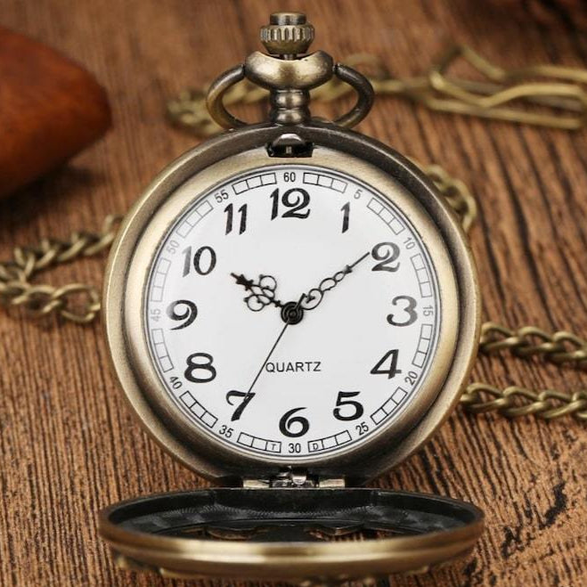 Antique bronze pocket watch with white face. Numbers indicated on face with black lettering. Gold chain on wood surface.