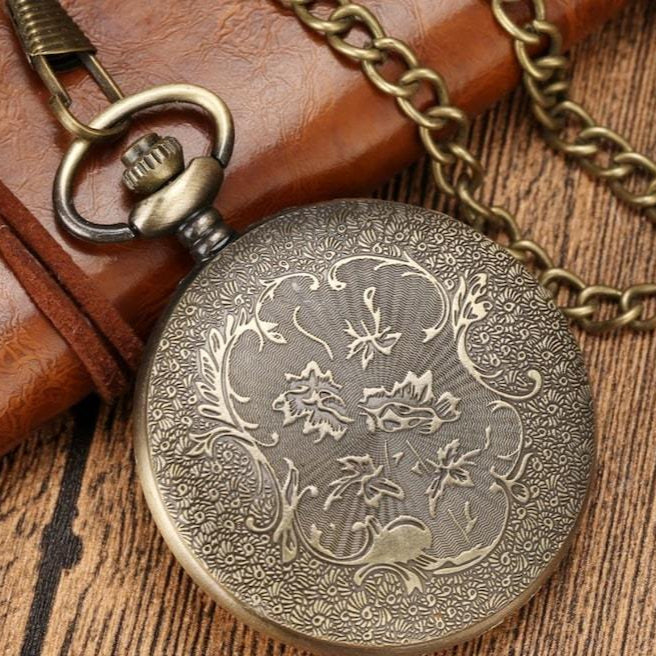 Close-up of an antique bronze pocket watch with a decorative floral design, attached to a matching chain, resting on a wooden surface and a brown leather item.