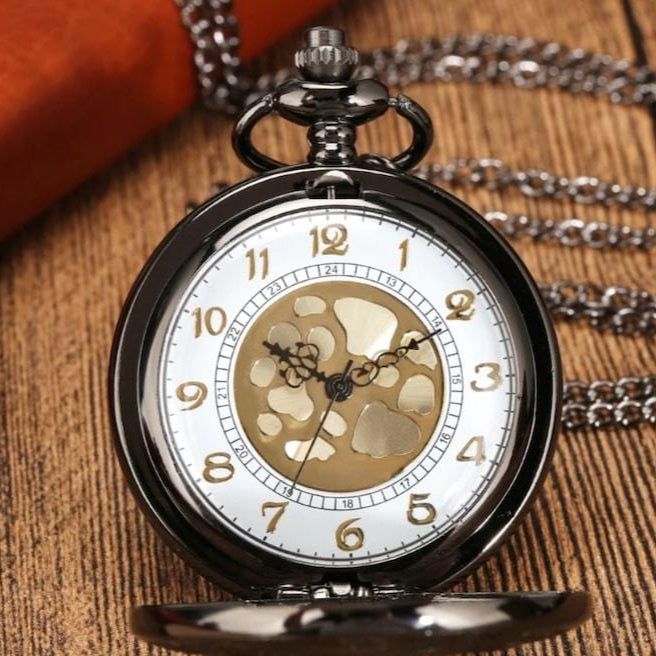Close-up of an open, black and gold mechanical pocket watch resting on a wood surface, with an orange wallet and silver chain in the background.