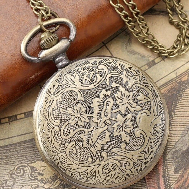 Close-up of an antique bronze pocket watch with an intricate floral design. It has a matching bronze chain and sits on a weathered map beside a brown leather object.
