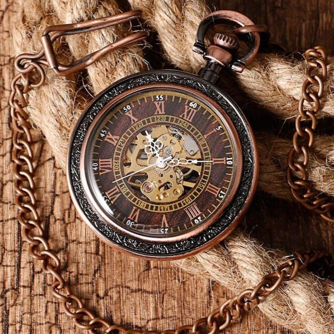 A copper-colored pocket watch with a chain on a wooden surface and rope.

