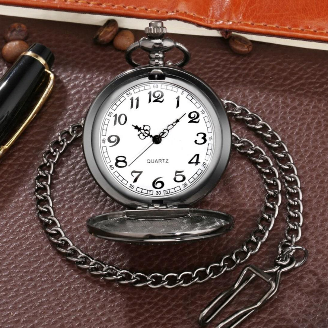 Close-up of a silver pocket watch with a chain, atop a brown leather surface. Pen and coffee beans are also visible.
