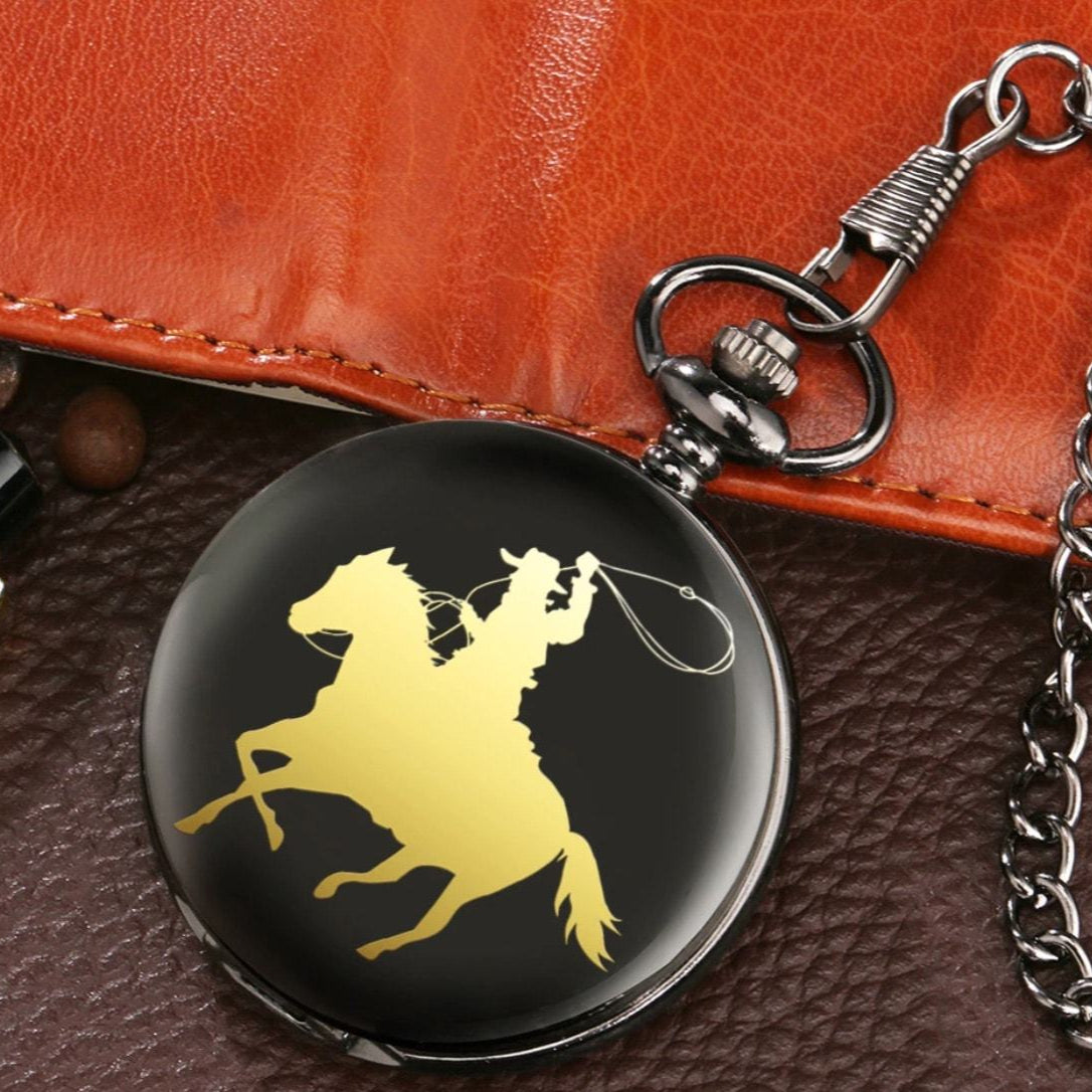 Close-up of a round, black pocket watch with a gold silhouette of a cowboy on a horse, atop a brown leather surface. The watch has a silver chain and clasp.