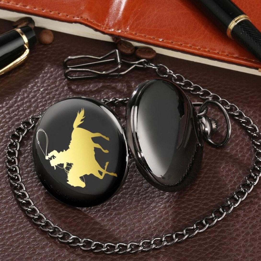 A black pocket watch with a gold rodeo design on a brown leather surface.
