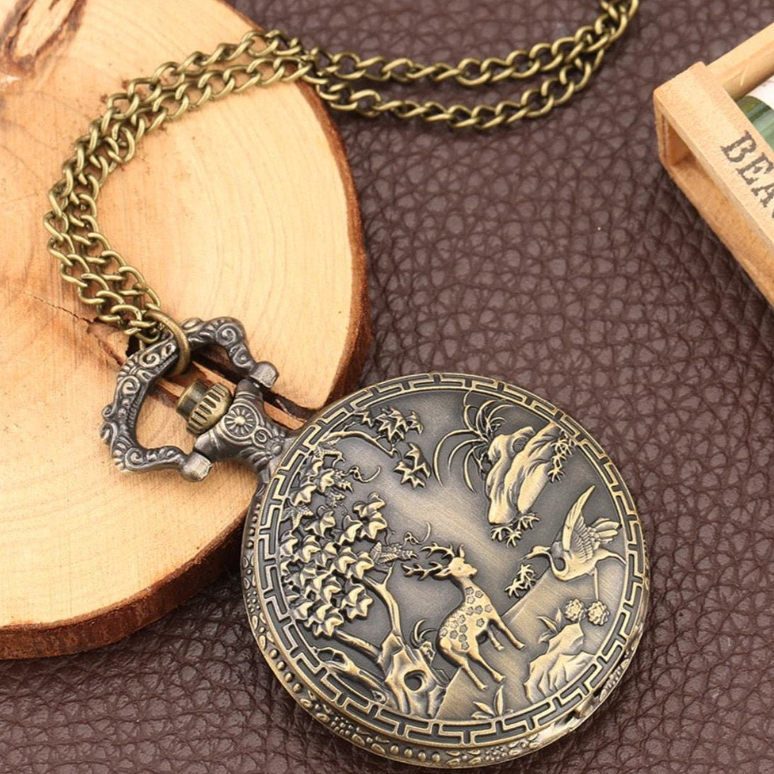 Antique bronze-toned pocket watch on a chain, displayed on a rustic wooden slice and textured brown surface. The watch features an intricate embossed design of a deer and birds.
