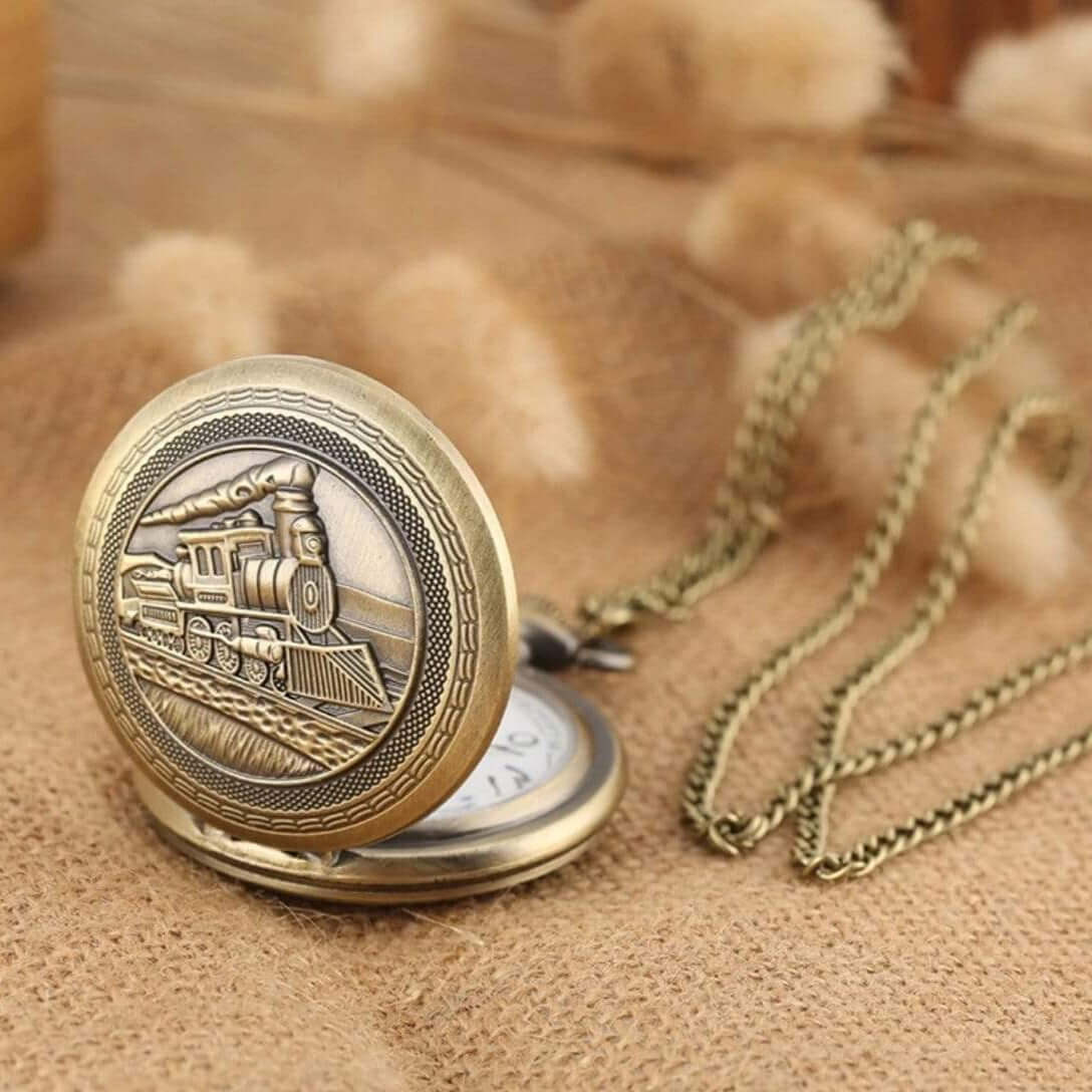 Close-up of an antique-style bronze pocket watch with a train design on the cover, accompanied by a bronze chain, on a tan burlap surface.
