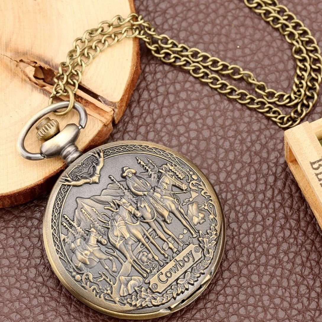 Antique bronze pocket watch with a cowboy theme, featuring a chain, set on a brown leather surface alongside a wooden accent.