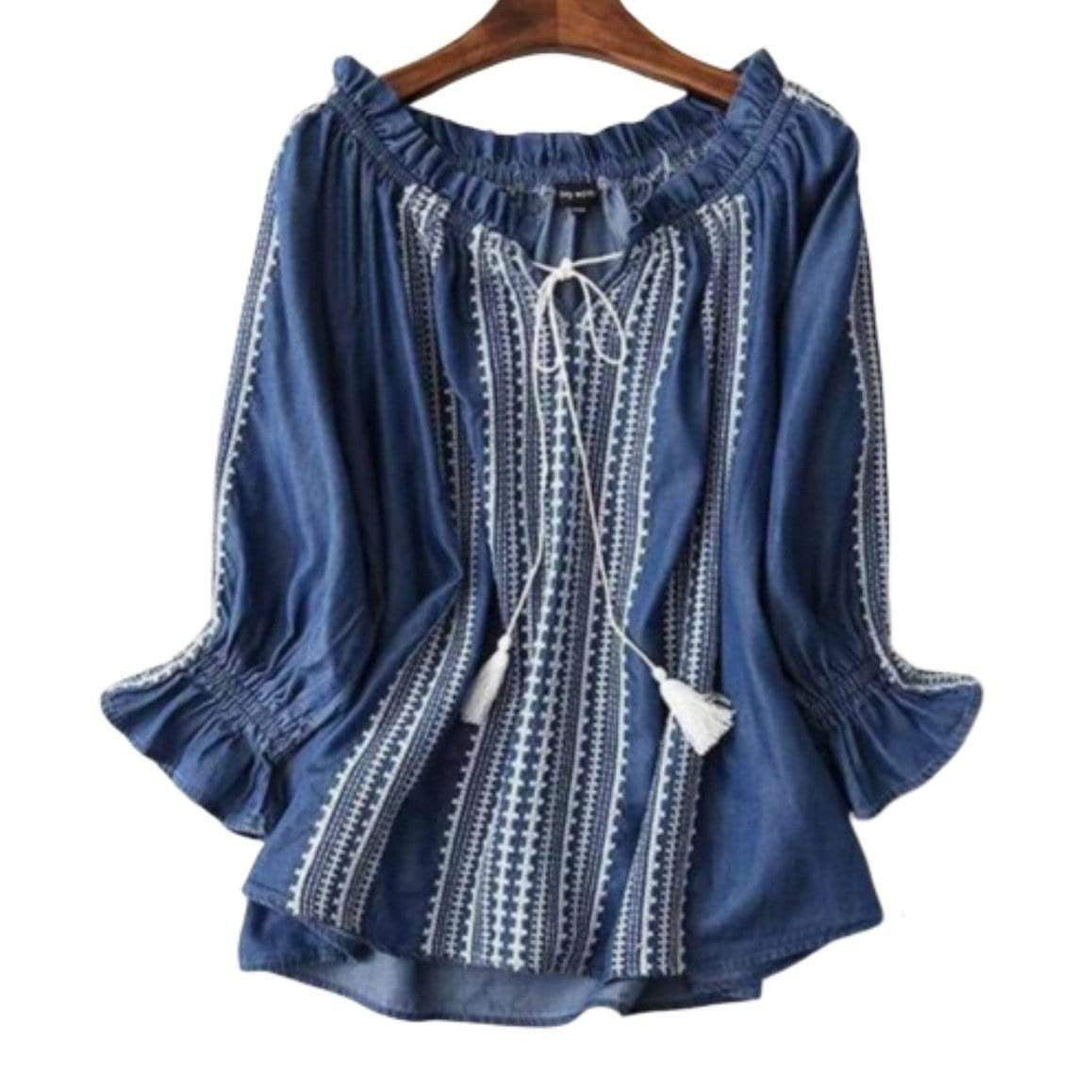 Blue peasant blouse with white embroidery, tassels, and ruffled neckline on wooden hanger.
