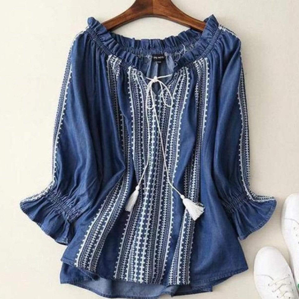 Blue denim blouse with white embroidery on a wooden hanger, paired with white sneakers.